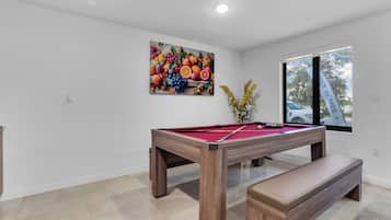 Una mesa de billar con un banco de madera, un cuadro con motivos de frutas en la pared y una ventana con vistas a un coche.