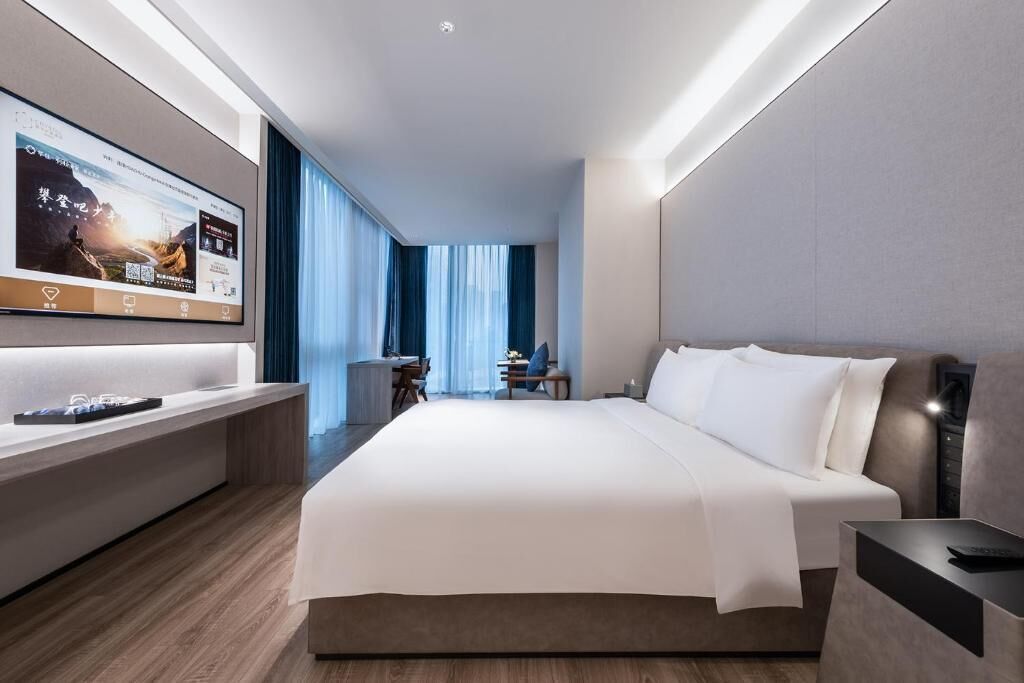 A modern hotel room with a large bed, a desk, a TV mounted on the wall, and a view of the outdoors through the window.