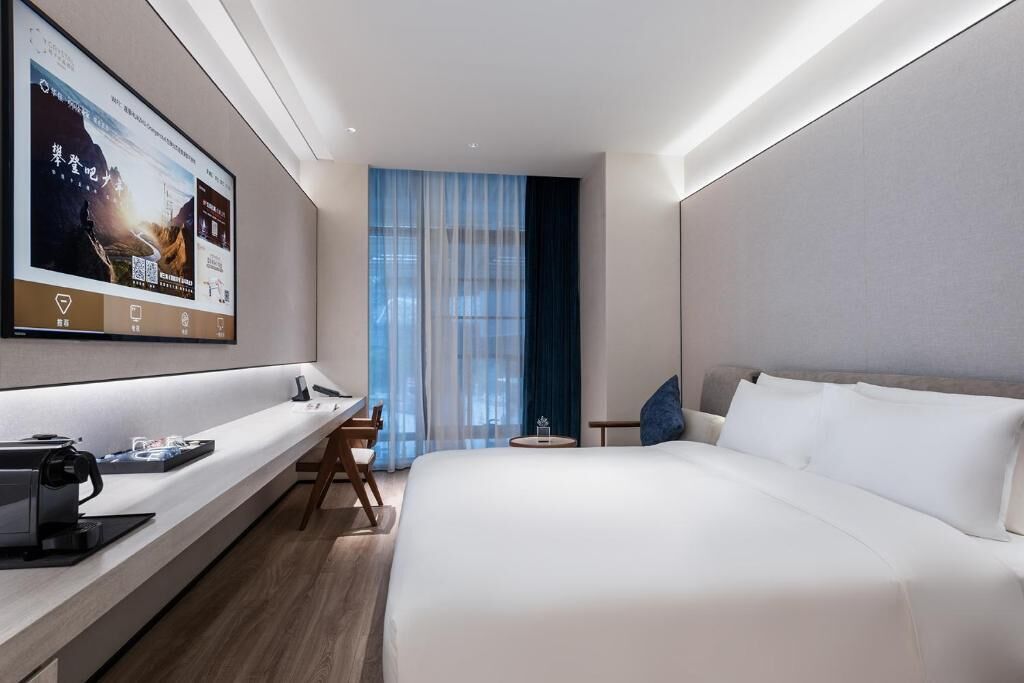 A modern hotel room with a large bed, a desk with a coffee machine, a TV mounted on the wall, and a view of the outdoors through a window.