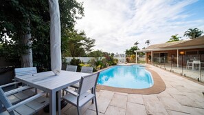 Une piscine avec un espace patio comprenant une table et des chaises, le tout entouré d’une clôture et de végétation.