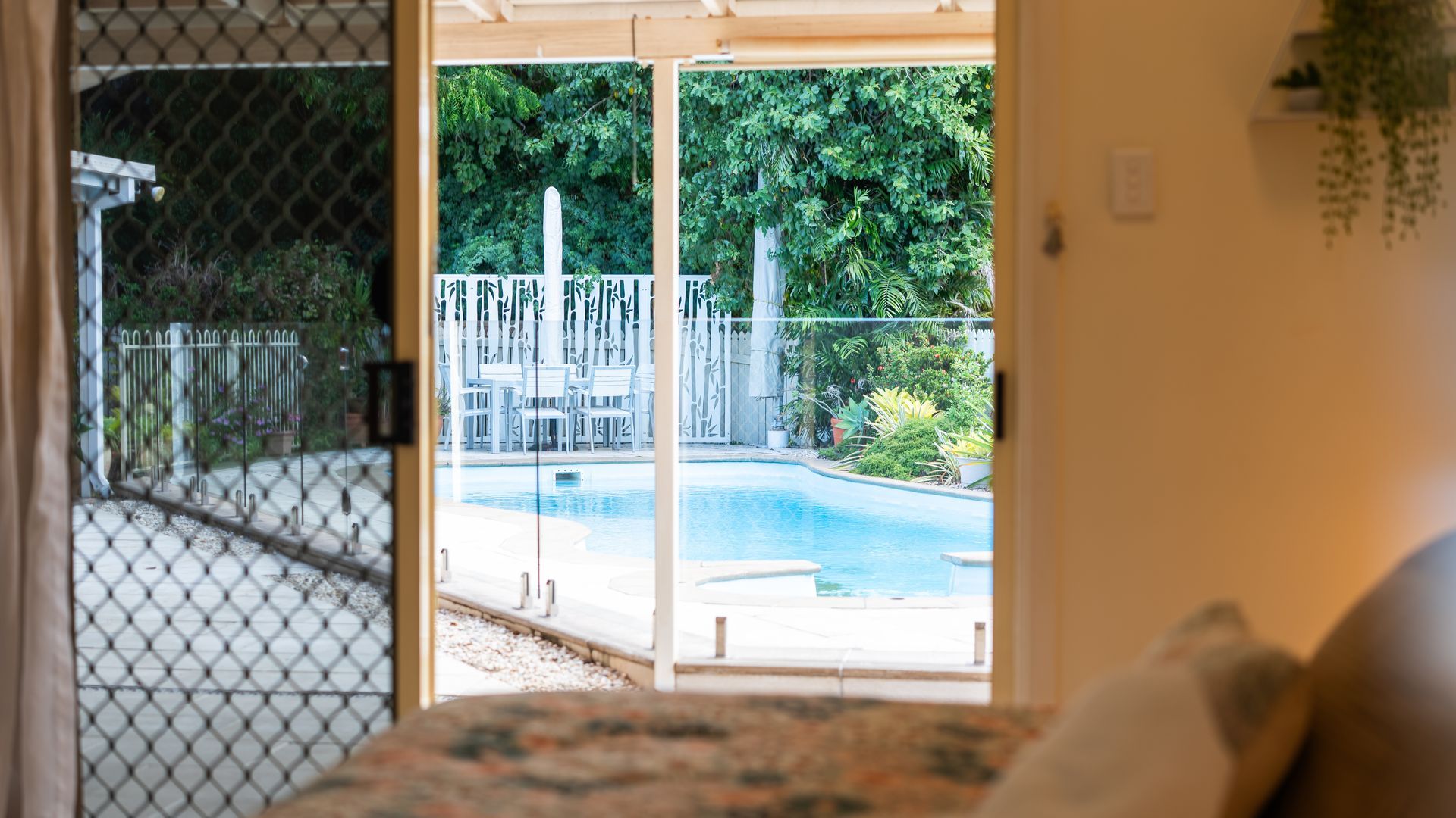 A view through a sliding glass door to a backyard with a swimming pool, surrounded by a fence and greenery.