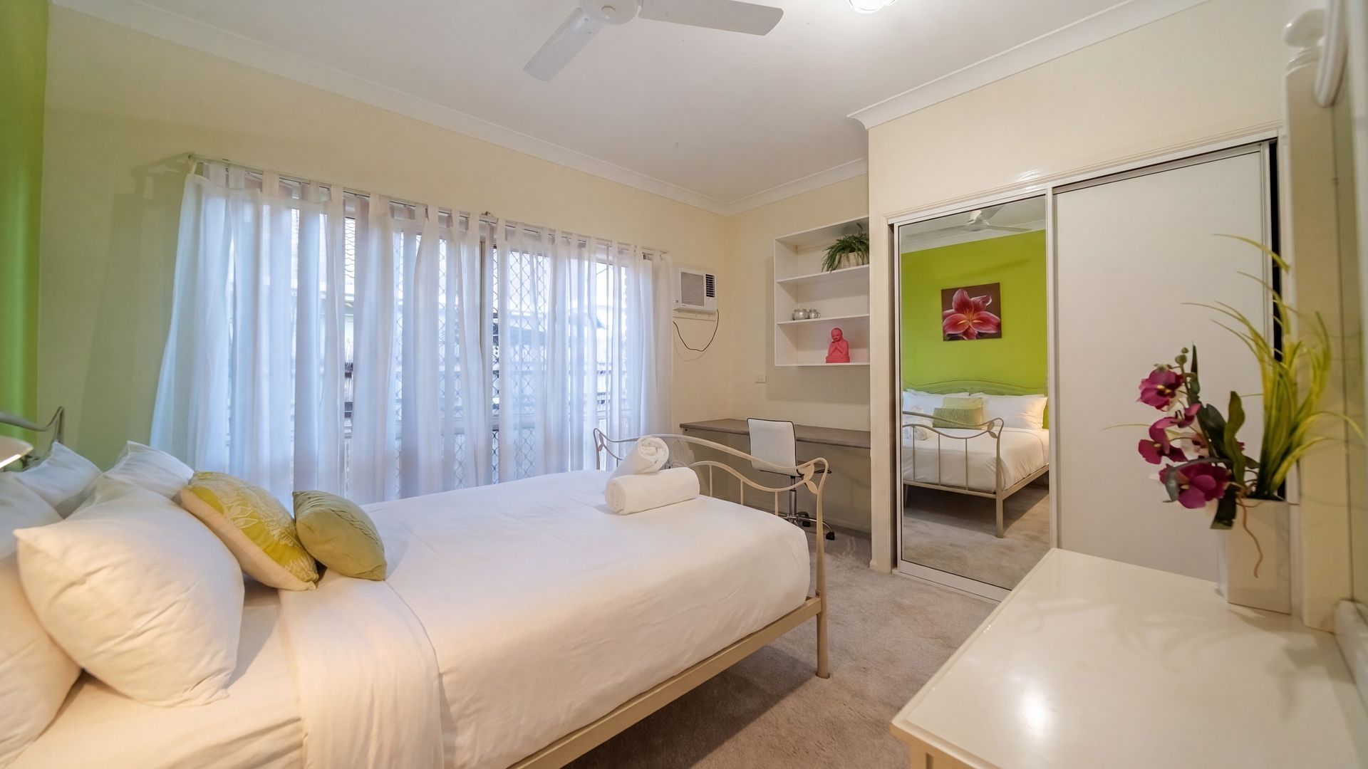 A hotel room with a bed, a desk, a chair, a mirror, and a plant.