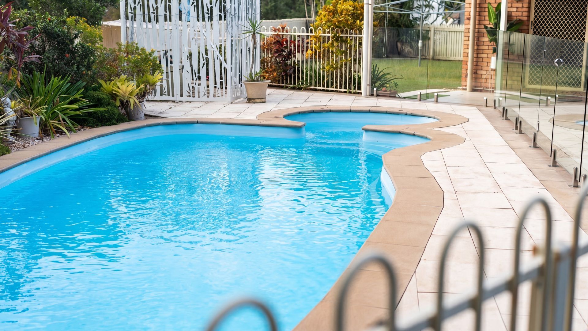 A swimming pool with a curved edge, surrounded by a tiled deck and a white decorative fence.