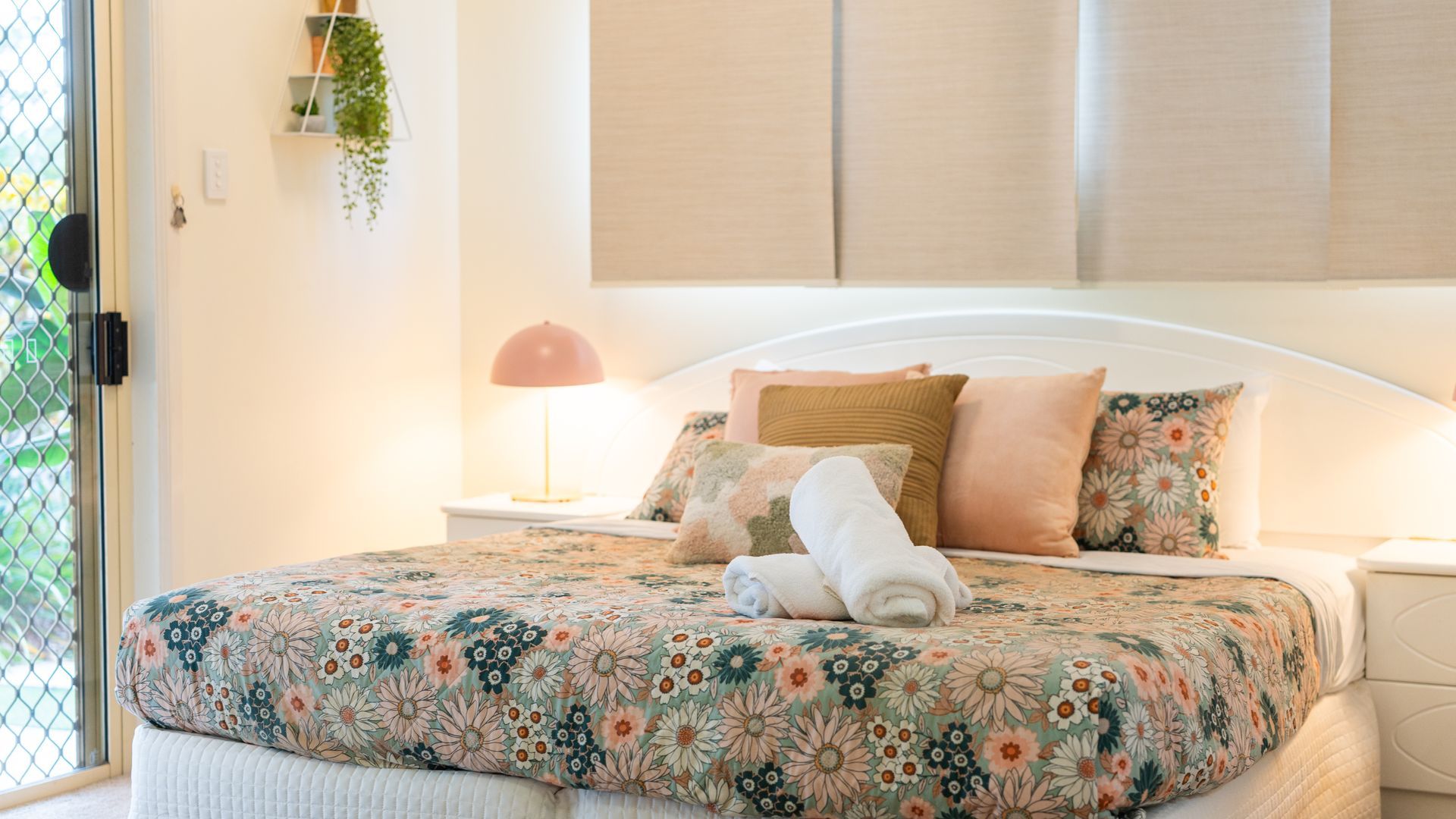 A neatly made bed with a floral bedspread, a bedside lamp, and a hanging plant.