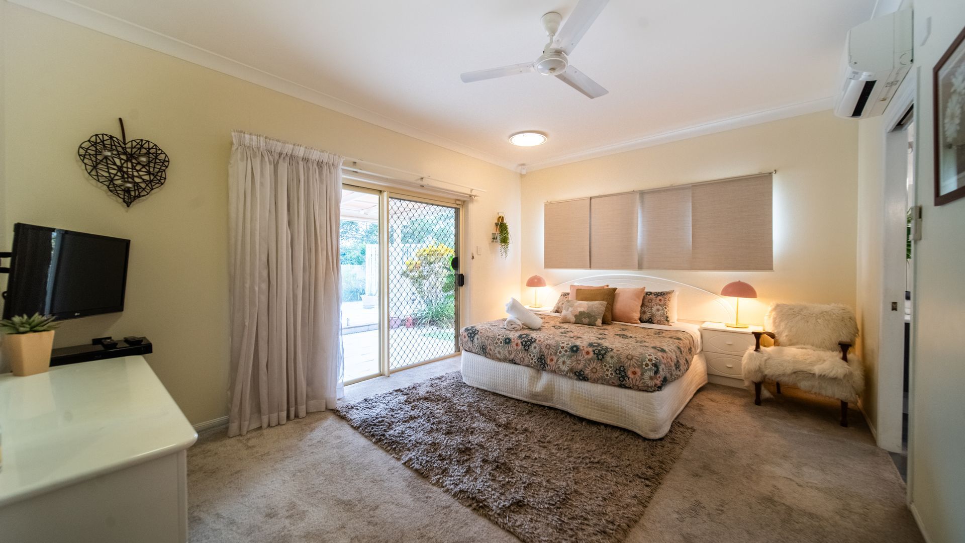 A bedroom with a bed, a desk, a TV, a ceiling fan, and a sliding glass door.