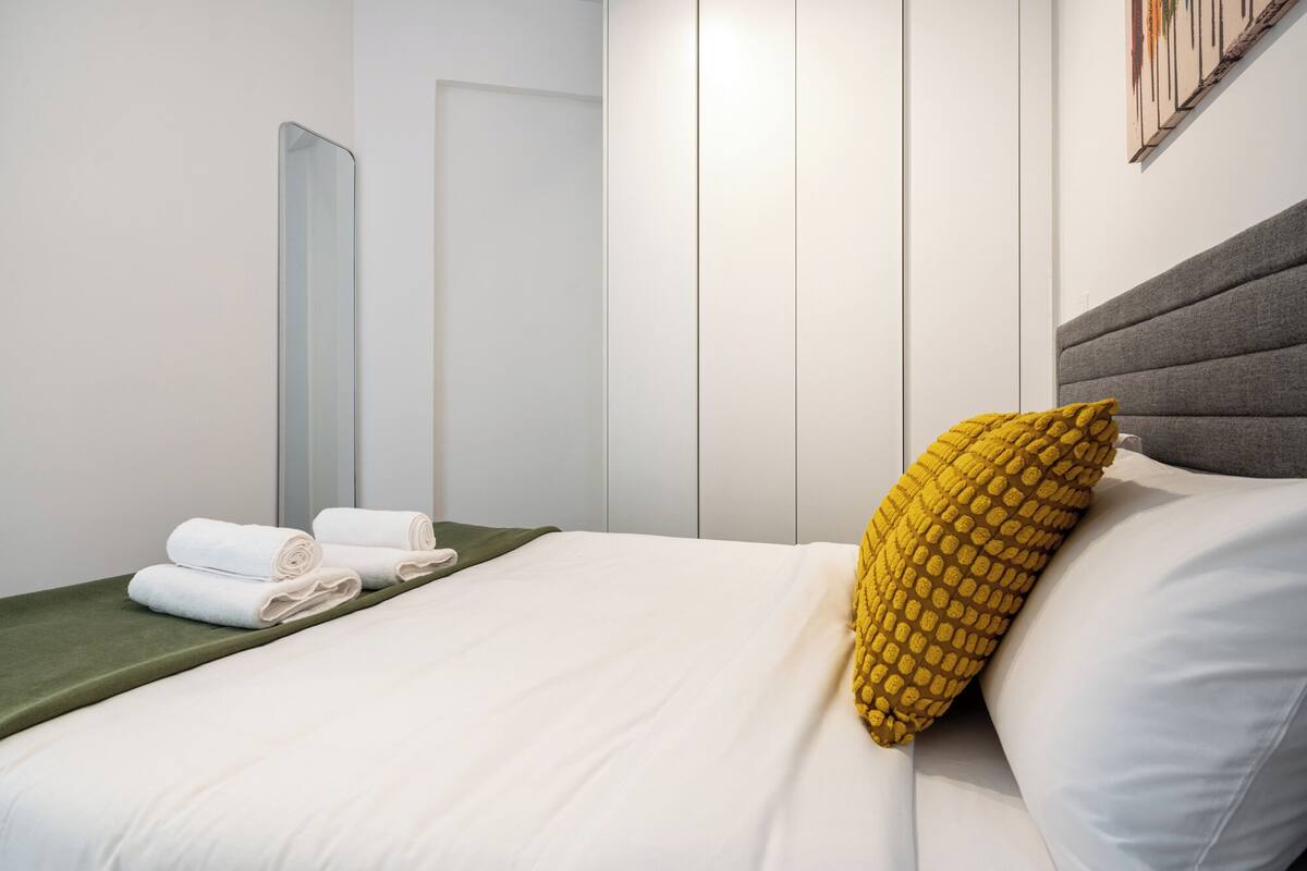 A hotel room with a bed, pillows, a green bedspread, a mirror, and a wall-mounted artwork.