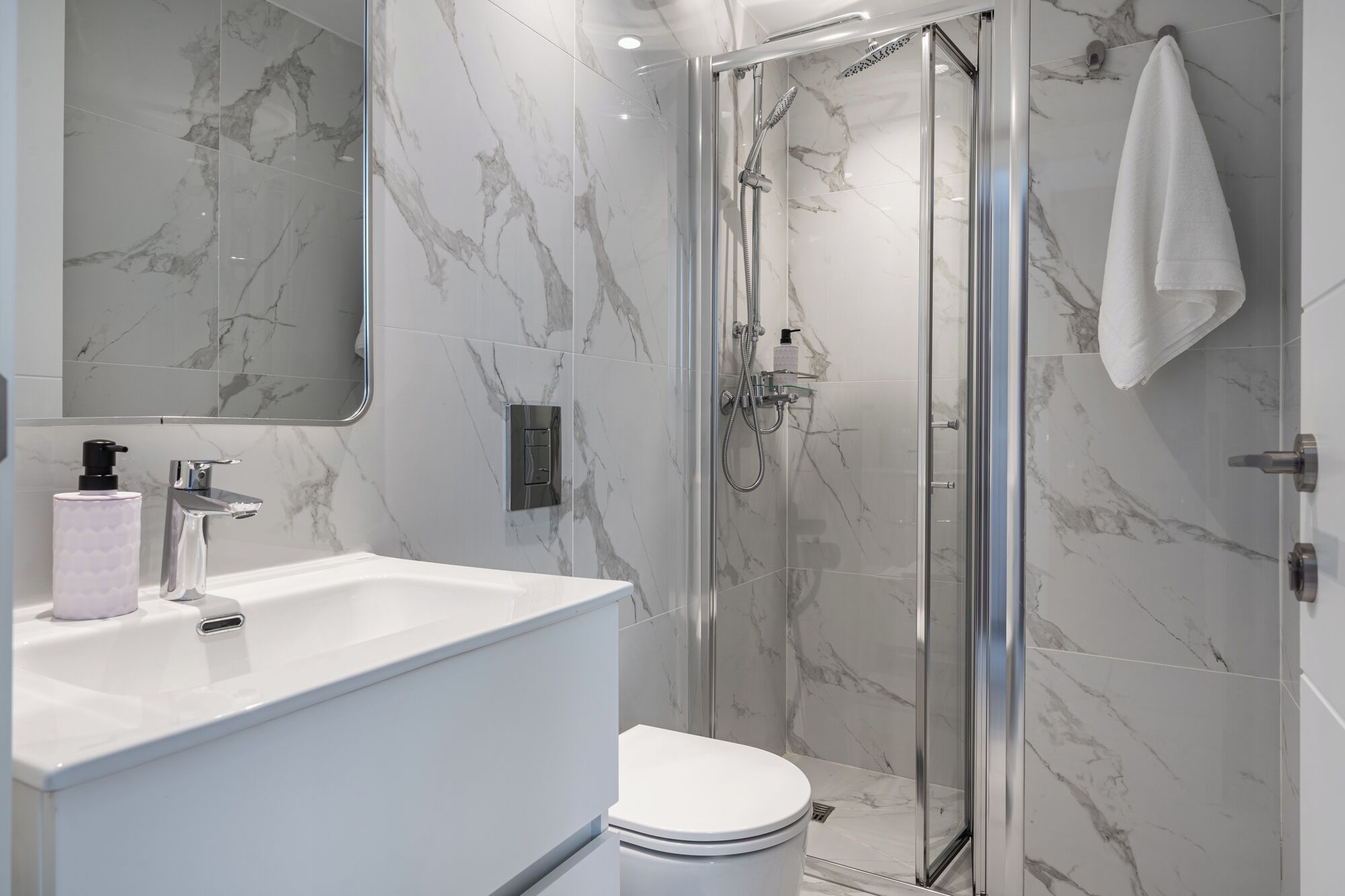 A modern bathroom with a glass-enclosed shower, a white toilet, a marble-tiled wall, and a large mirror.