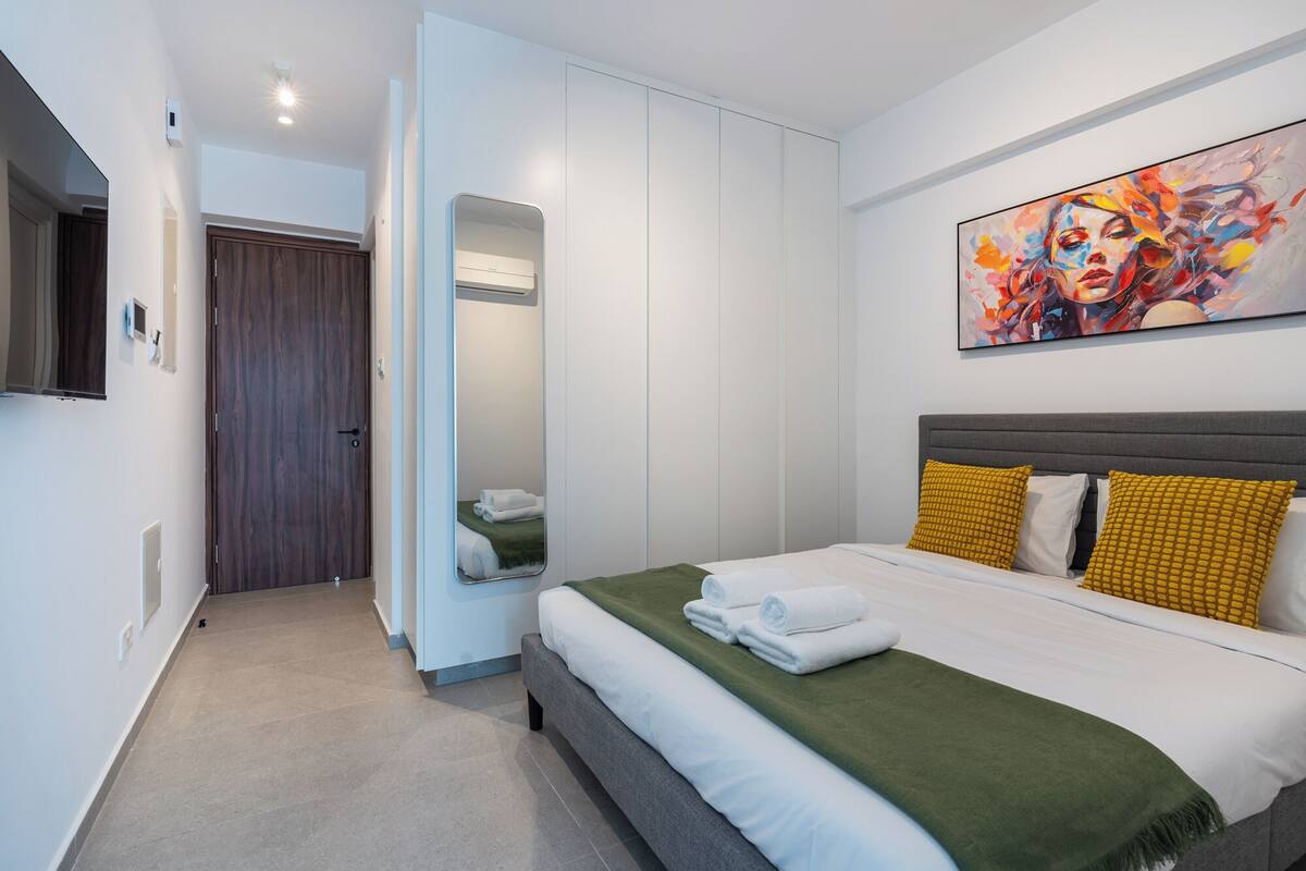 A modern hotel room with a bed, a large mirror, a painting on the wall, and a door leading to another room.