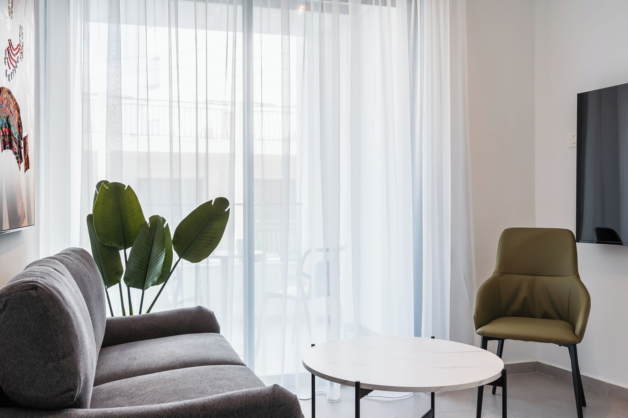 A modern living room with a grey sofa, a round white table, a green armchair, and a large window with sheer curtains.