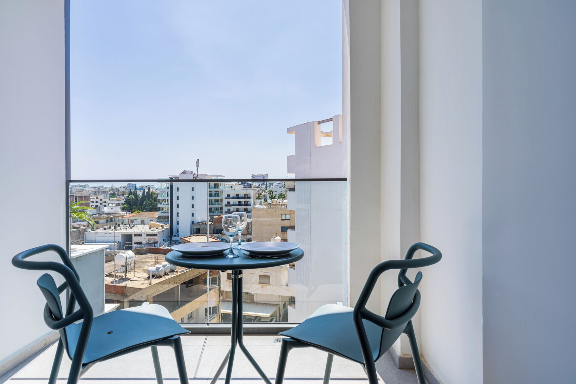 A balcony with a table and chairs, a clear view of the cityscape, and a modern building in the background.