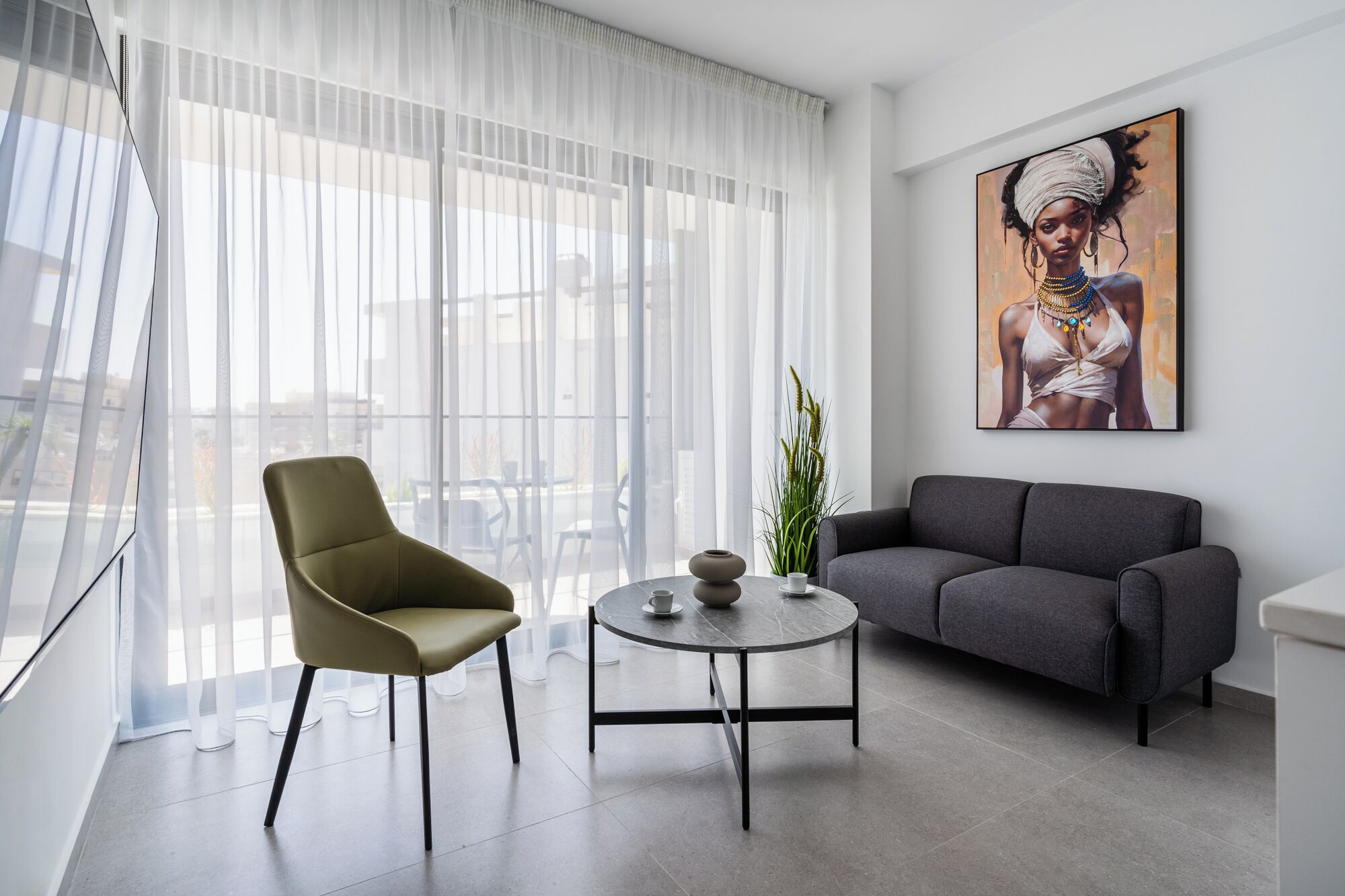 En moderne stue med sofa, stol, rundt bord og et stort vindue med lette gardiner.