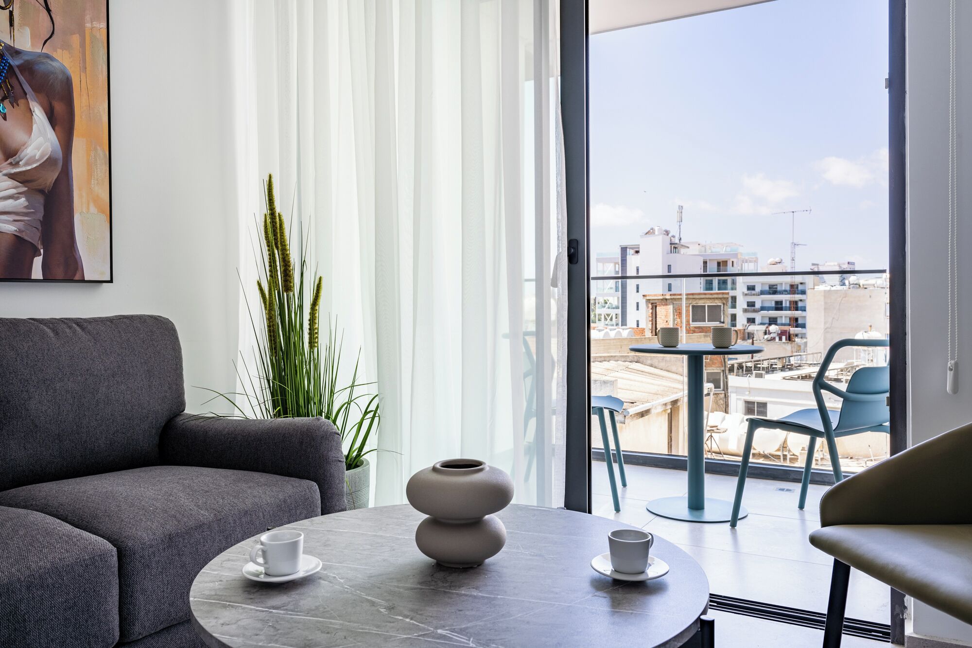 A modern living room with a grey sofa, a round table with two white cups, a large window with a view of buildings, and a potted plant.
