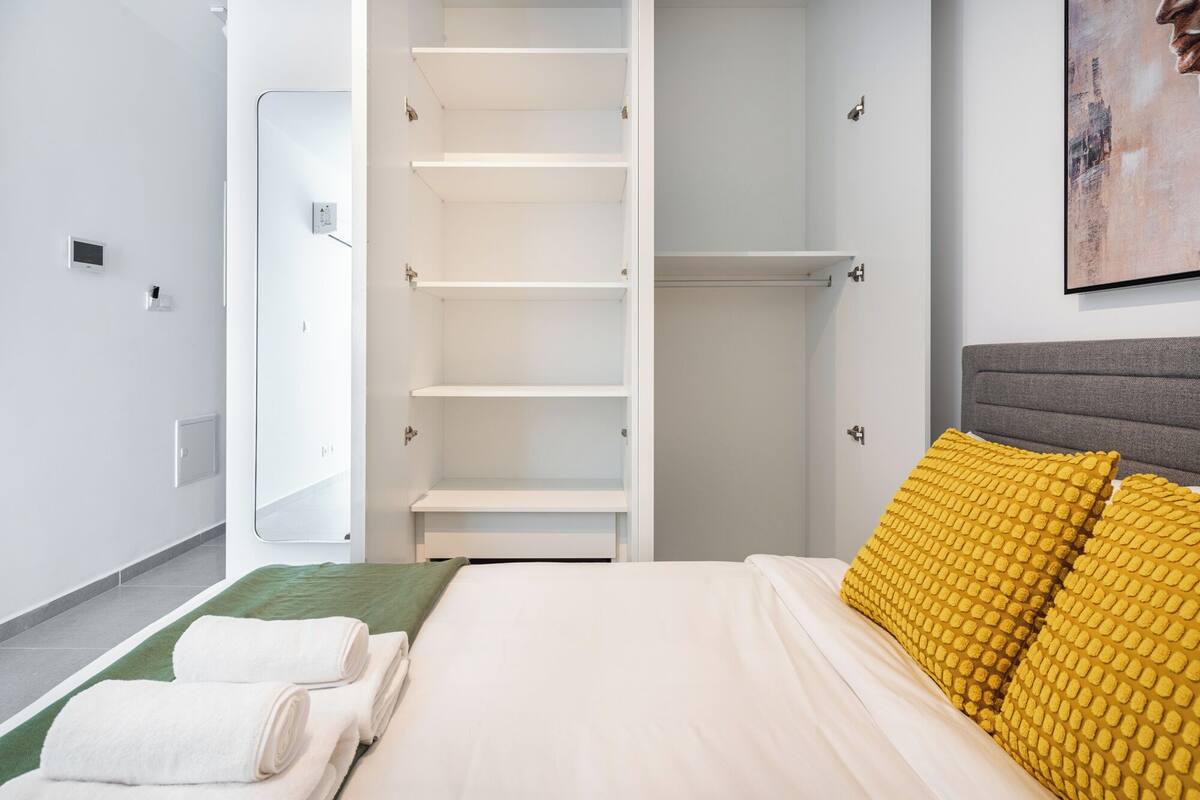 A hotel room with a bed, a green area rug, a white wall, a white wardrobe with open doors, and a painting on the wall.