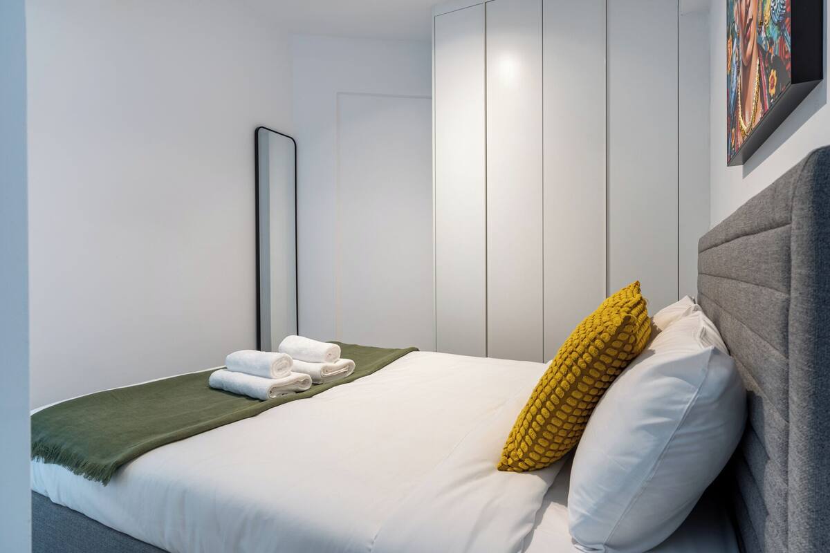 A hotel room with a bed, a green throw blanket, white pillows, a yellow pillow, a grey headboard, and a painting on the wall.