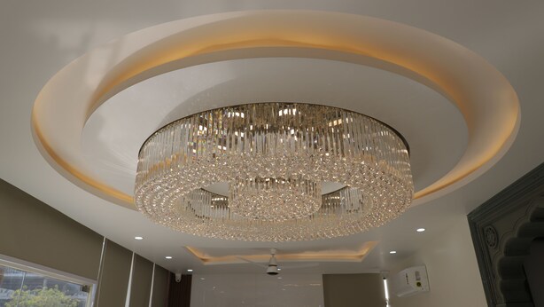 A modern interior with a large, ornate chandelier, recessed ceiling lighting, and a view of the outdoors through a window. - Krishneya Dham (Mathura)