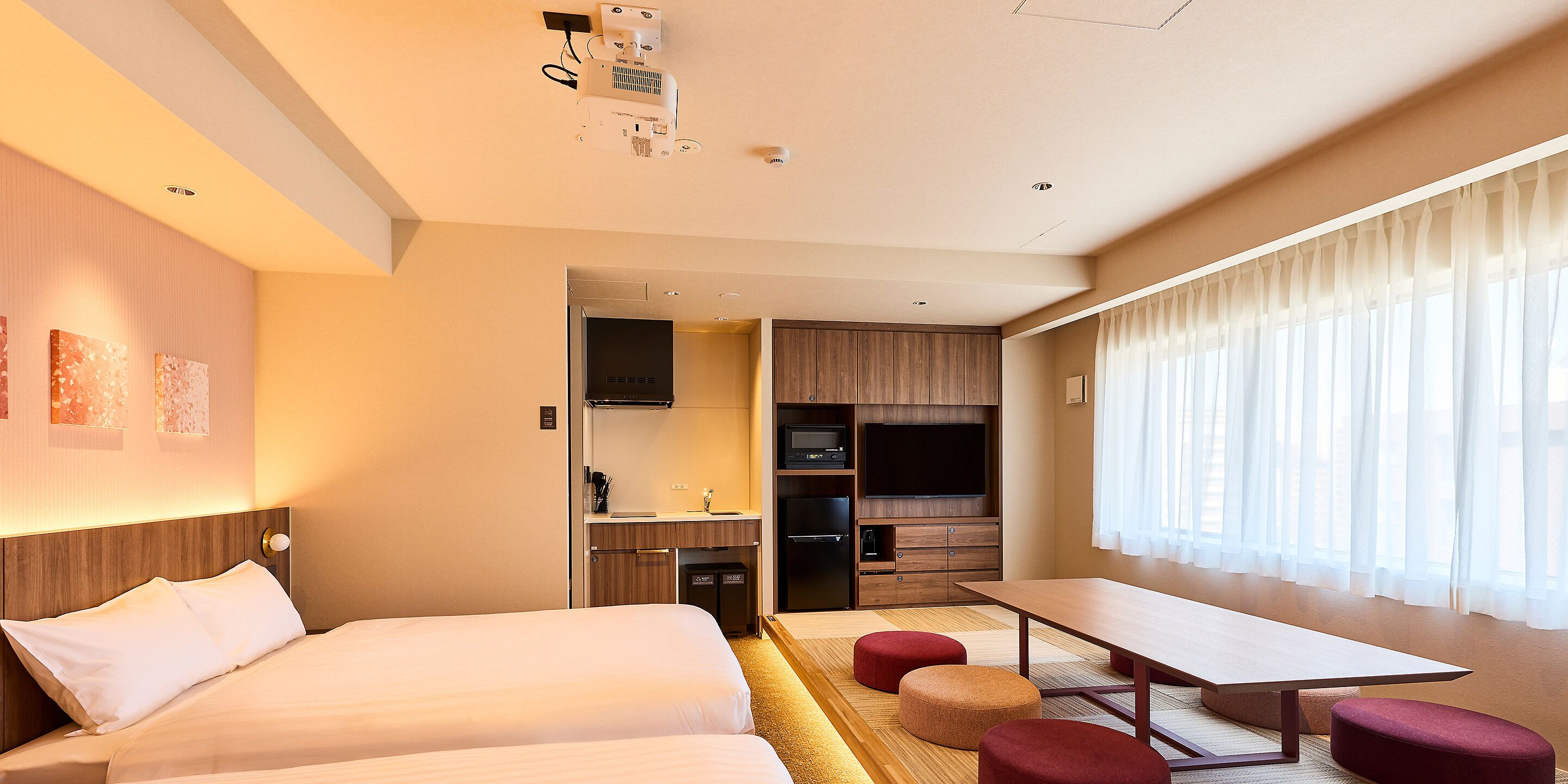 A hotel room with a bed, a desk, a kitchenette, and a large window with curtains.