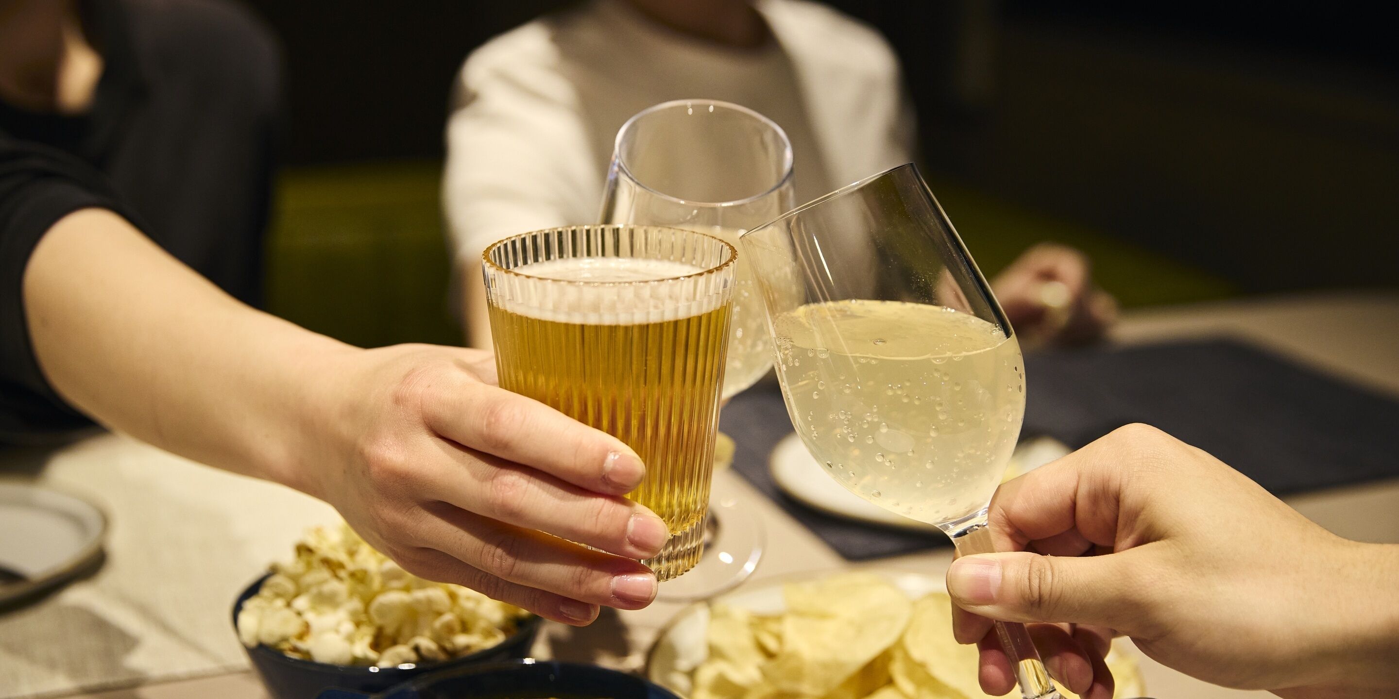 Two hands holding glasses of beer and wine, with a bowl of popcorn and a plate of chips on the table.
