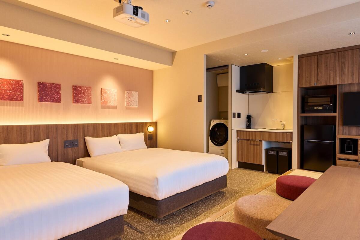 Habitación de hotel con dos camas, televisor, kitchenette y lavadora.