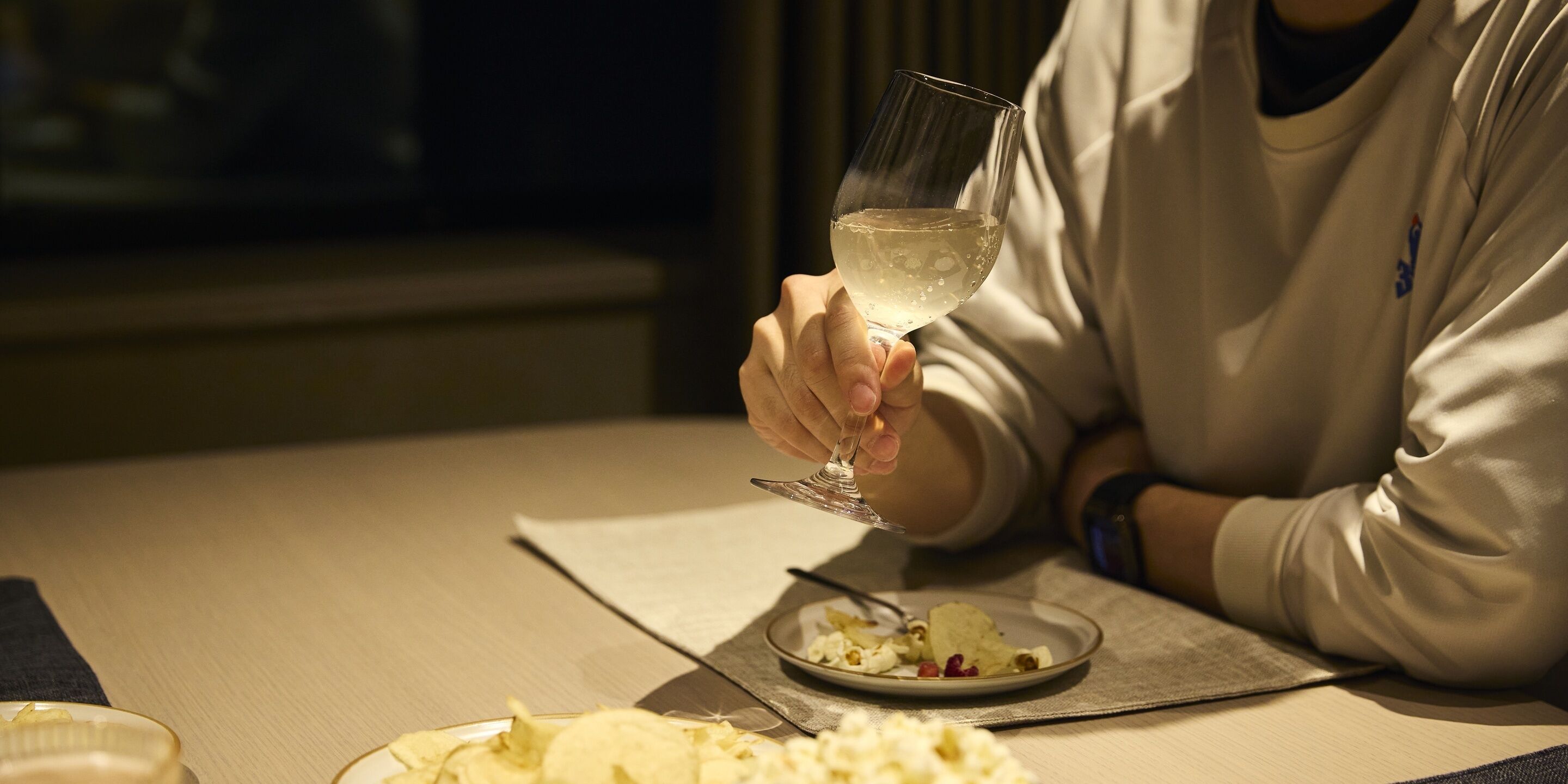 Una persona sosteniendo una copa de vino blanco en una mesa con un plato de comida y una servilleta.