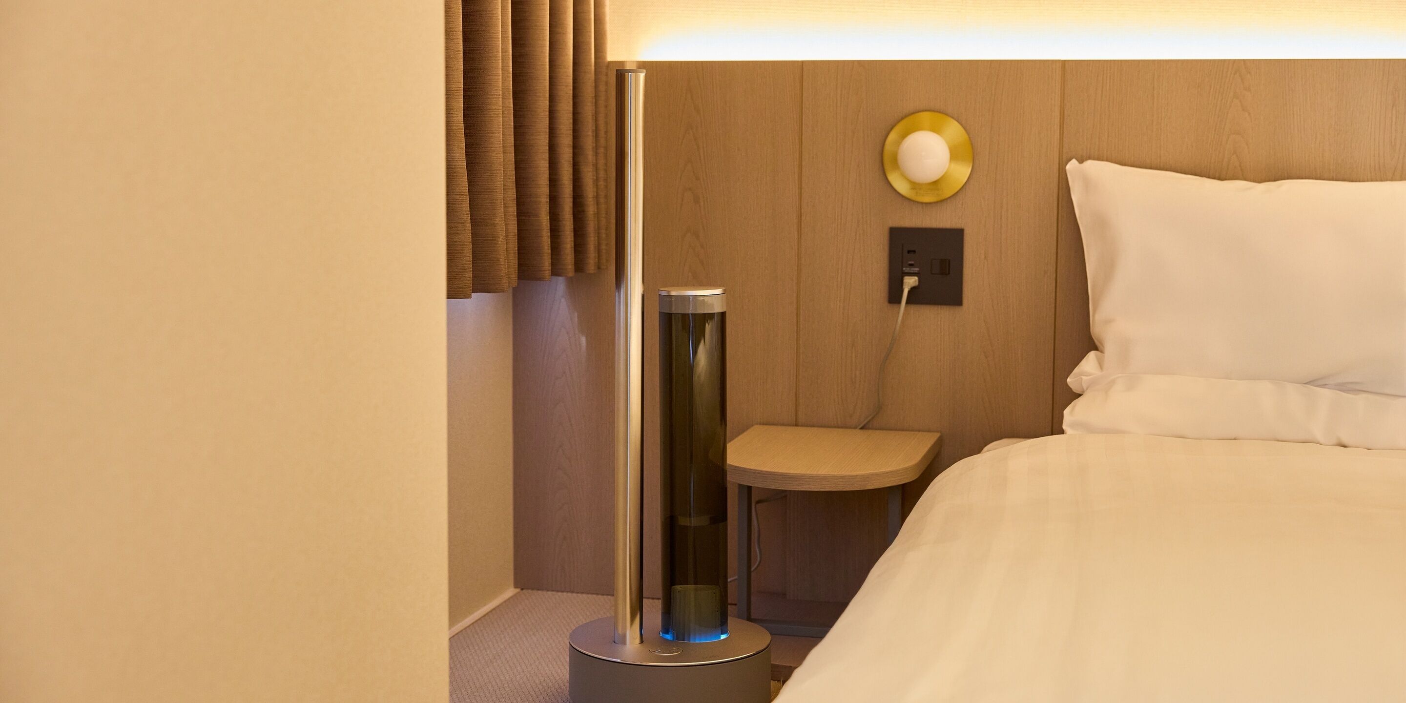 A hotel room with a bed, a bedside table, a lamp, a wall-mounted light, and a power outlet.