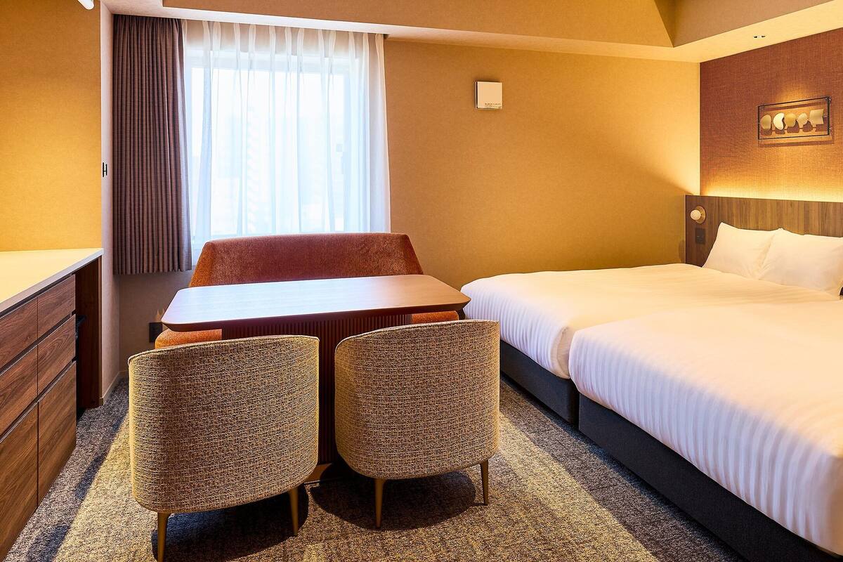 Una habitación de hotel con cama, escritorio, dos sillas, ventana con cortinas y una lámpara fijada a la pared.