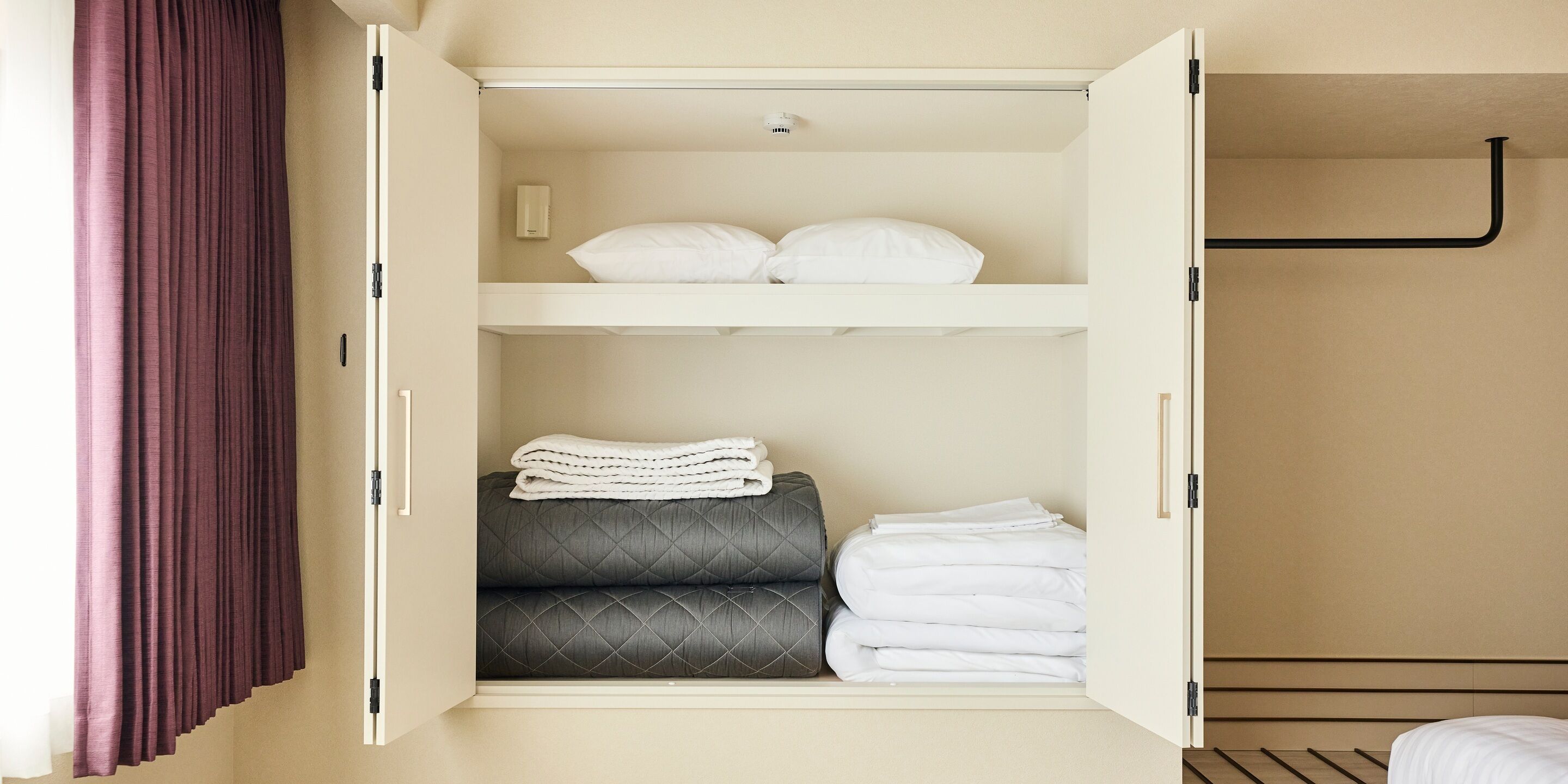 A hotel room with a wardrobe containing folded towels, a quilted grey sofa, and a bed with a black headboard.
