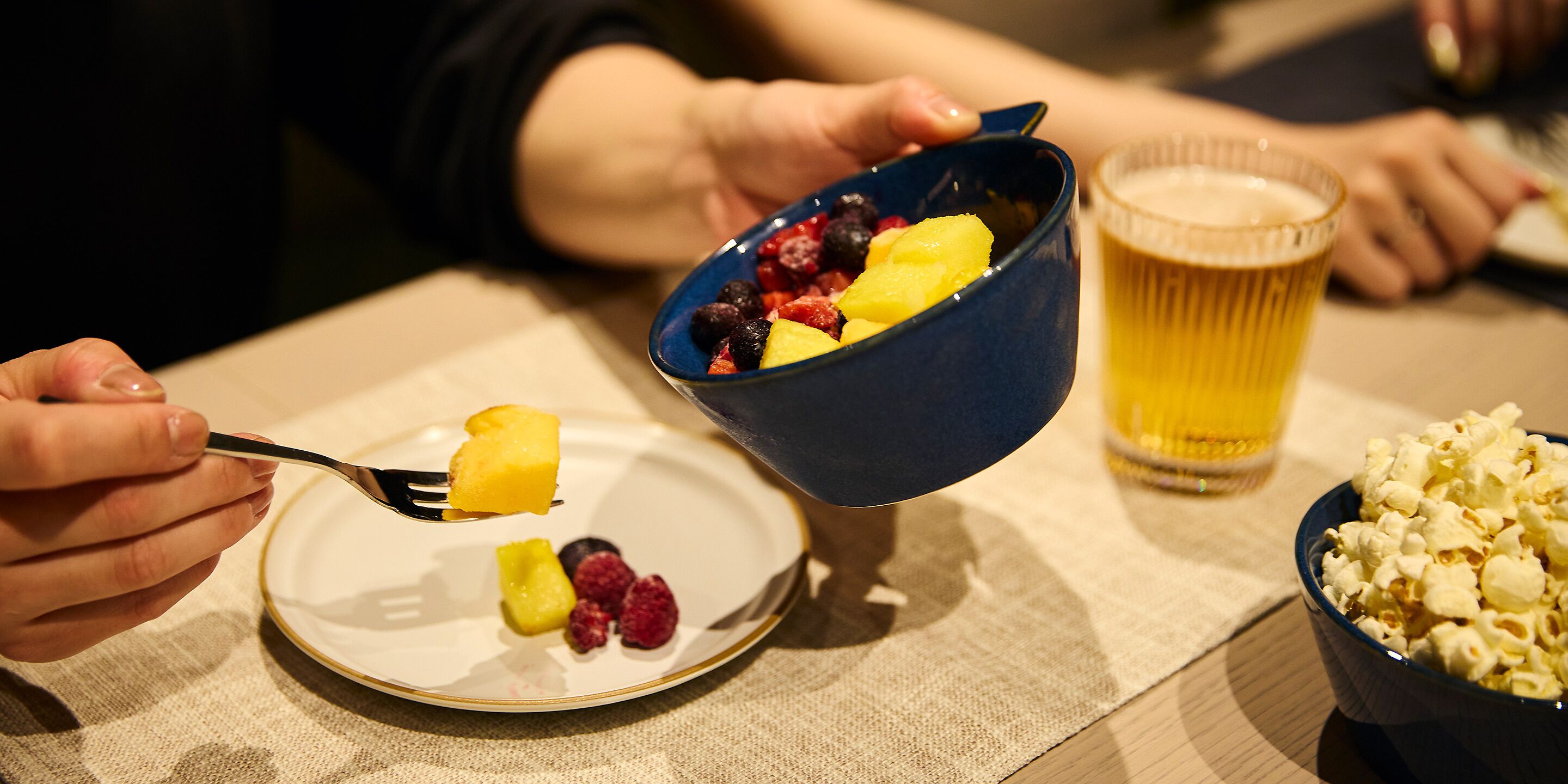A dining table with a bowl of fruit, a glass of beer, and a bowl of popcorn.