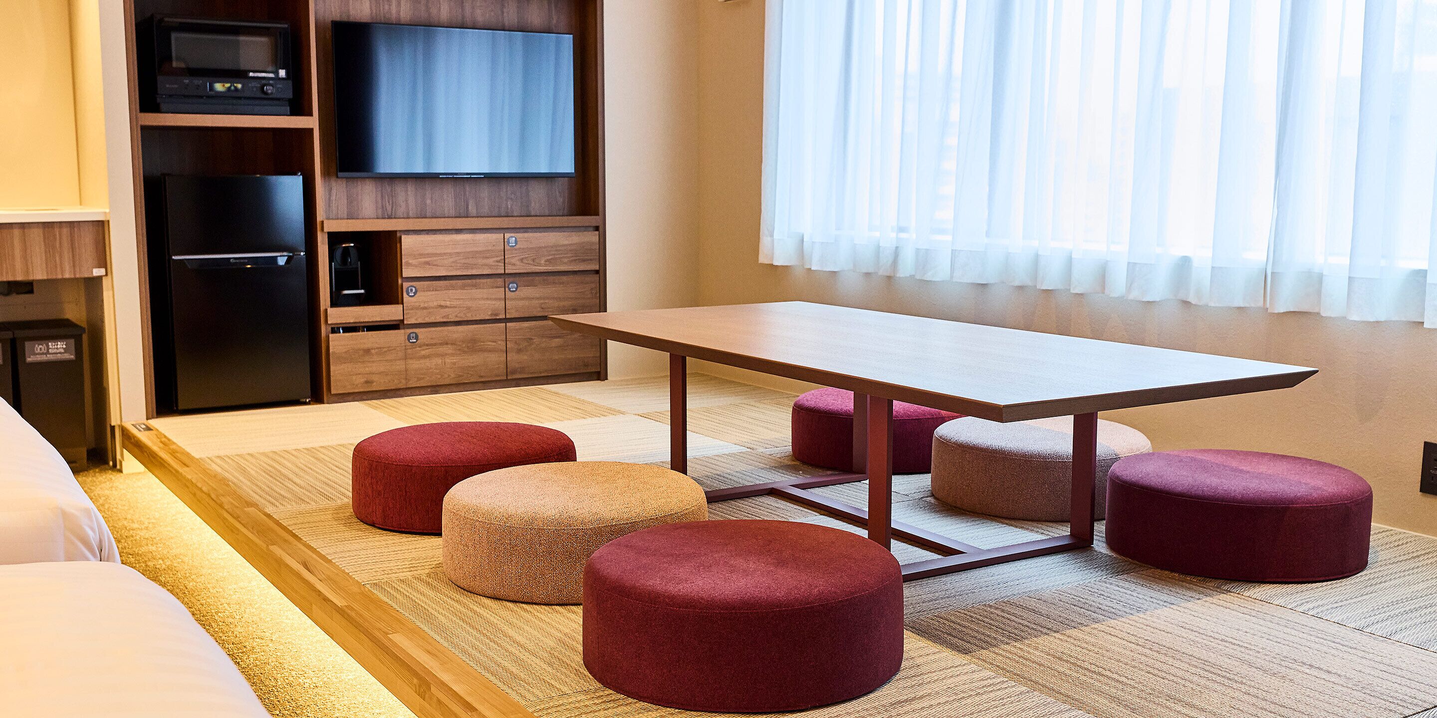 A hotel room with a wooden table, four ottomans, a television, and a minibar.