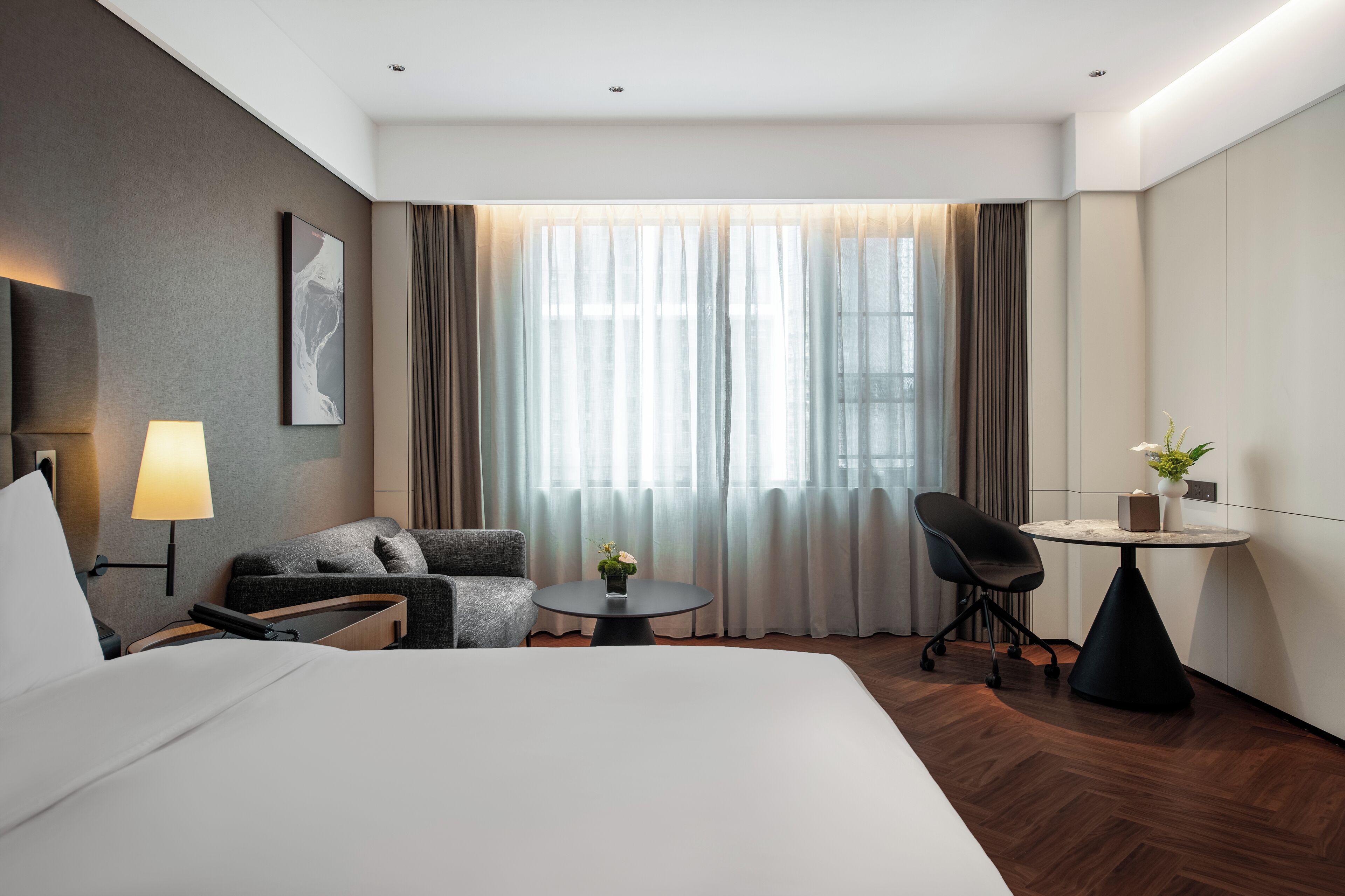 Une chambre d’hôtel avec un lit, un bureau, une chaise, une lampe, une petite table avec une plante et une fenêtre avec des rideaux.
