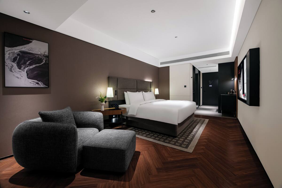 Una habitación de hotel moderna con una cama grande, un espacio de descanso con una silla y un reposapiés, una mesita de noche con lámpara, y un cuadro abstracto de gran tamaño en la pared.