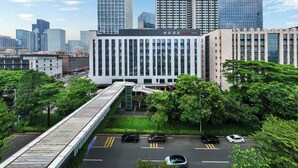 A modern building with a prominent entrance, surrounded by lush greenery and a pedestrian bridge.