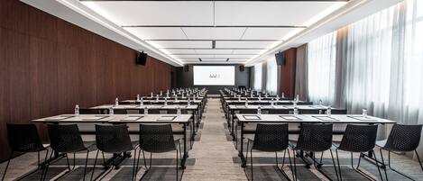 Un salón de conferencias con filas de mesas y sillas, una pantalla de proyección y un ventanal grande.