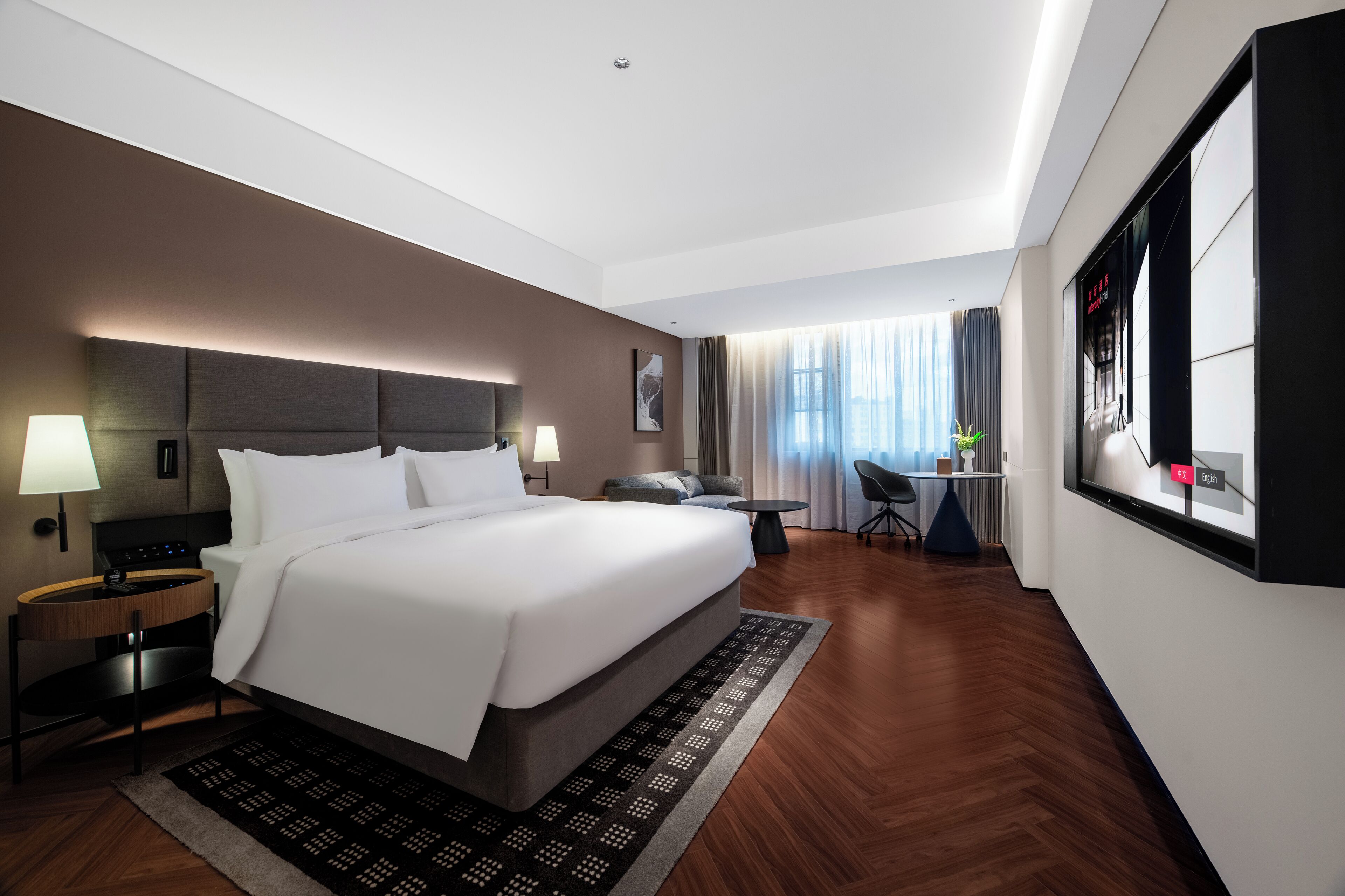 Quarto de hotel moderno com uma cama grande, mesas de cabeceira, escrivaninha e vista para a cidade.