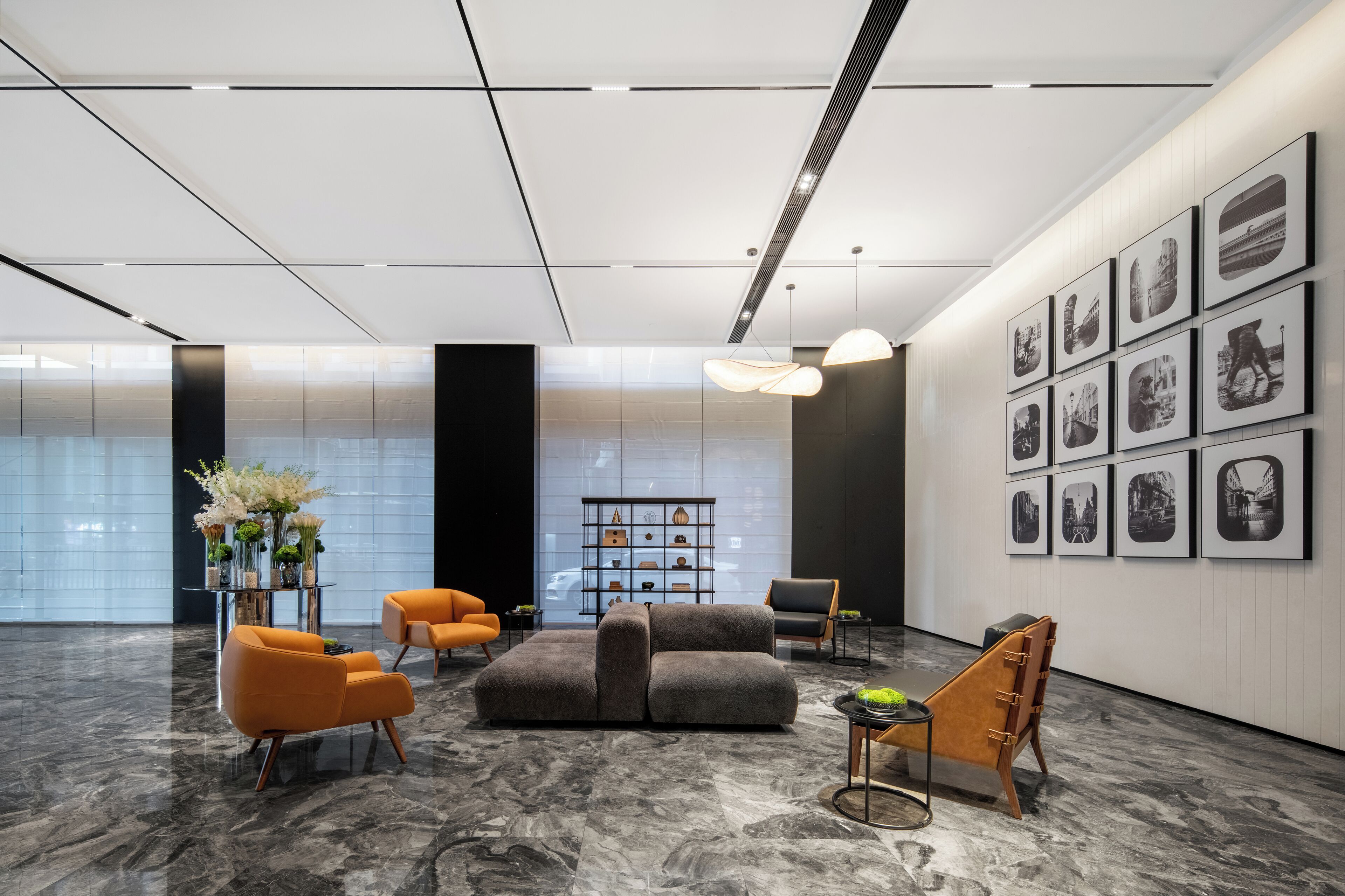 A modern lobby with a grey sofa, orange chairs, a black bookshelf, and framed pictures on the wall.
