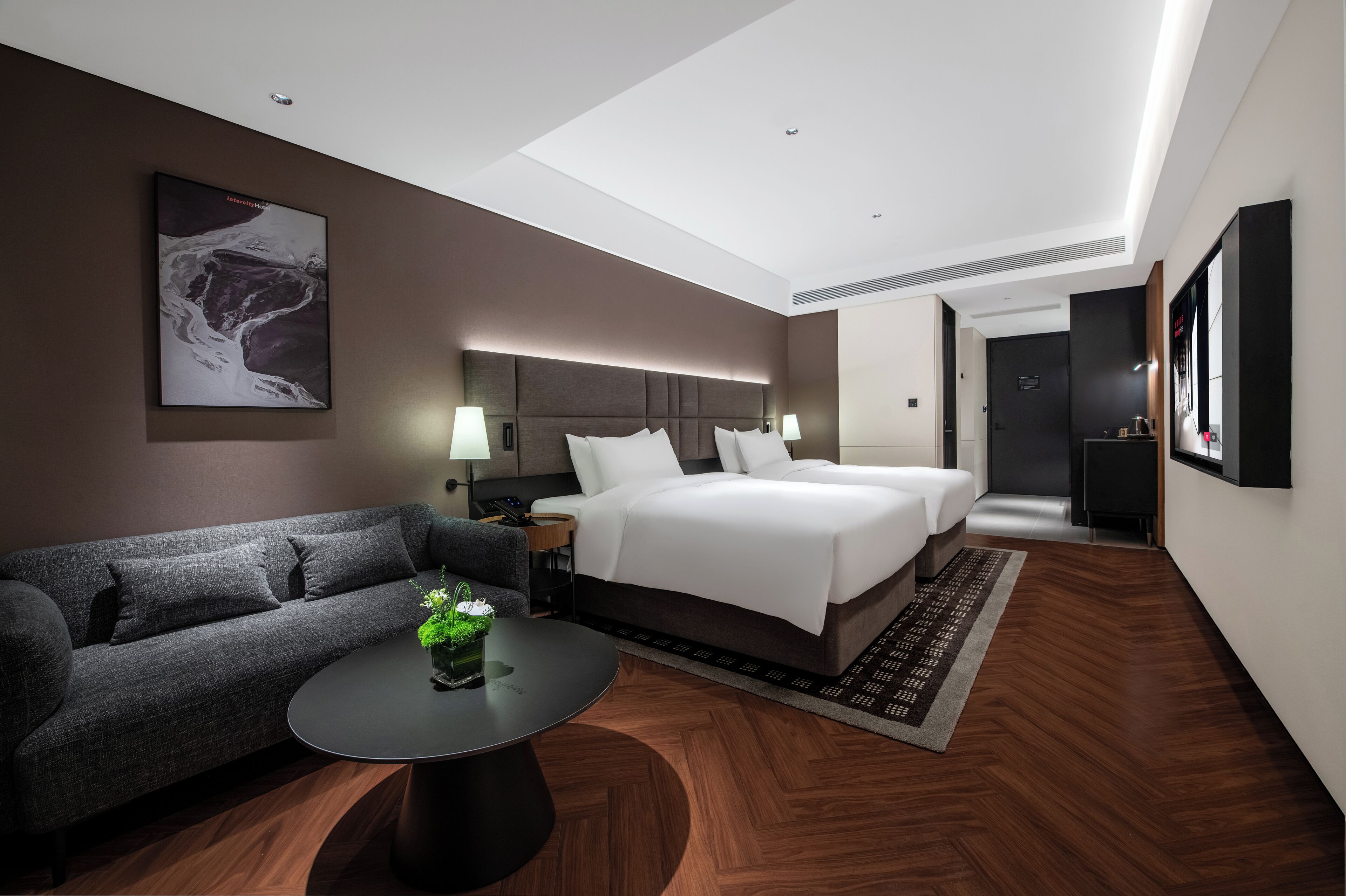 A modern hotel room with a large bed, a sofa, a small table with a plant, and a painting on the wall.