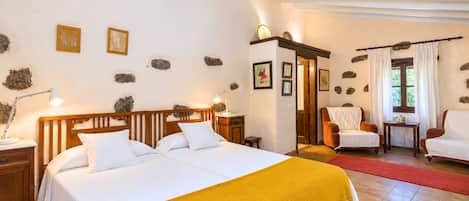 Un dormitorio con una cama doble, dos mesitas de noche, un buró, una silla, una pequeña mesa y una ventana con cortinas.
