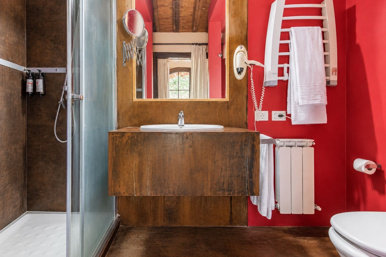 Een badkamer met een houten wastafelmeubel, een glazen douchecabine en een rode wand.