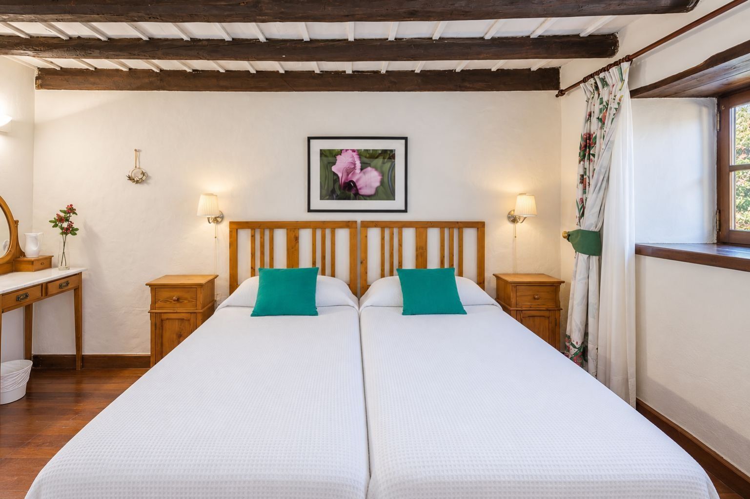 Cama doble con dos almohadas, cabecero de madera, lámparas de noche, un cuadro en la pared y una ventana con cortinas.