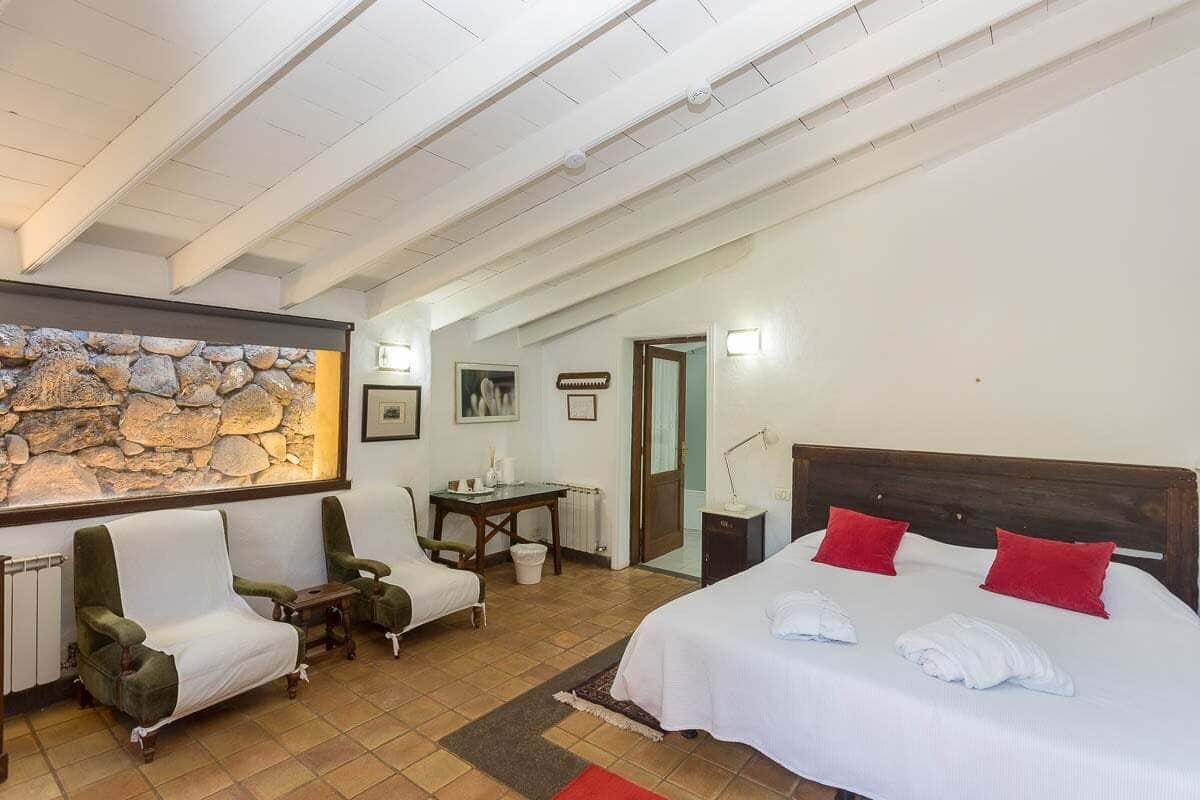 Una habitación de hotel con una cama, dos sillones, una mesita, una pared de piedra y una puerta que da a otra habitación.