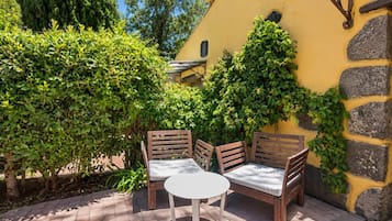 Un área de estar al aire libre con una mesa y dos sillas, rodeada de follaje y una pared amarilla.