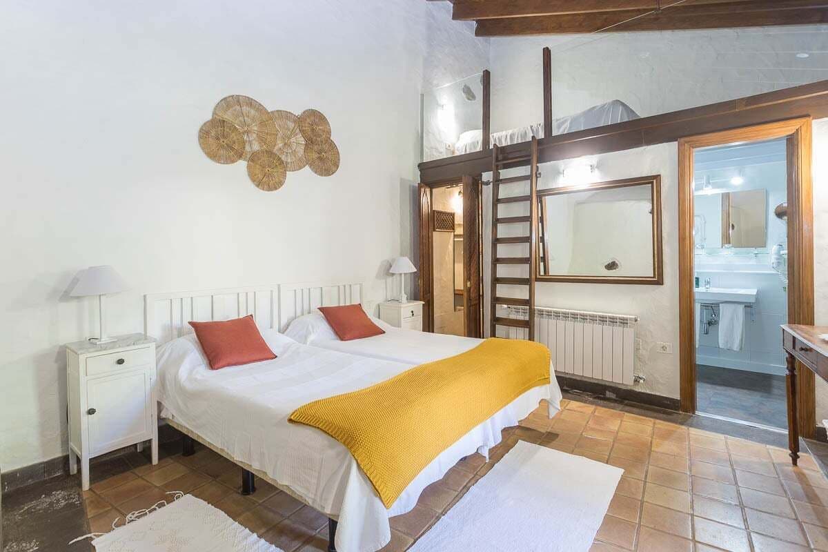Un dormitorio con una cama, dos mesitas de noche, un espejo, una escalera y un baño visible a través de una puerta abierta.