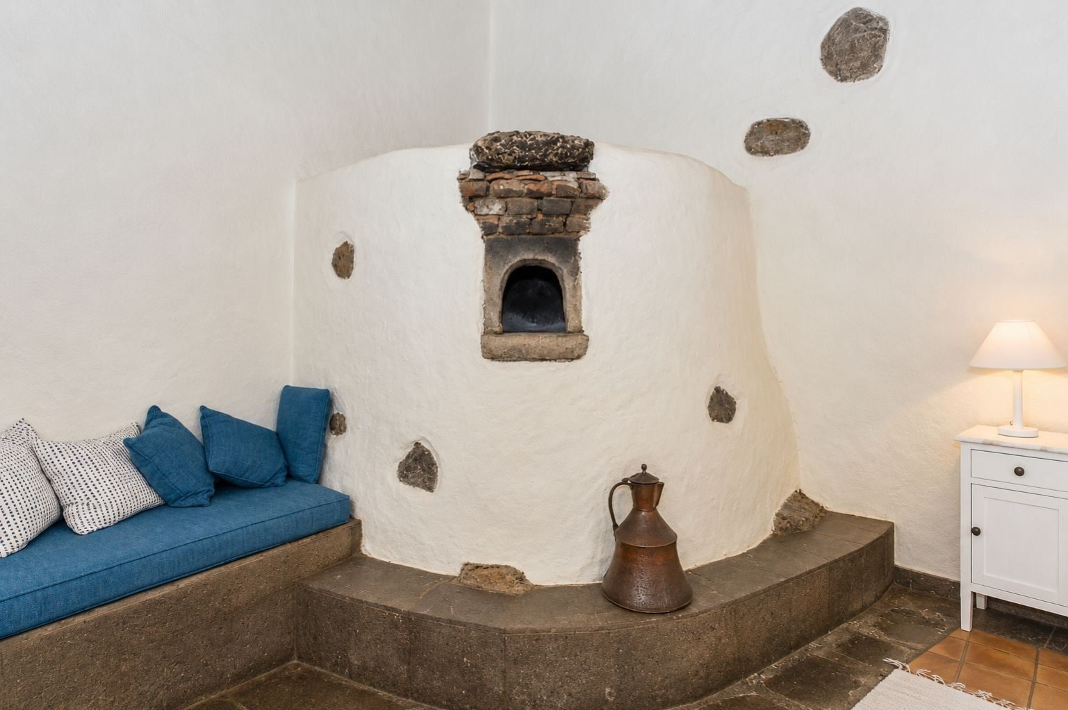 Una habitación con pared blanca con textura, una chimenea de piedra, un sofá azul con cojines estampados, una jarrón de metal y una mesita de noche blanca con una lámpara.