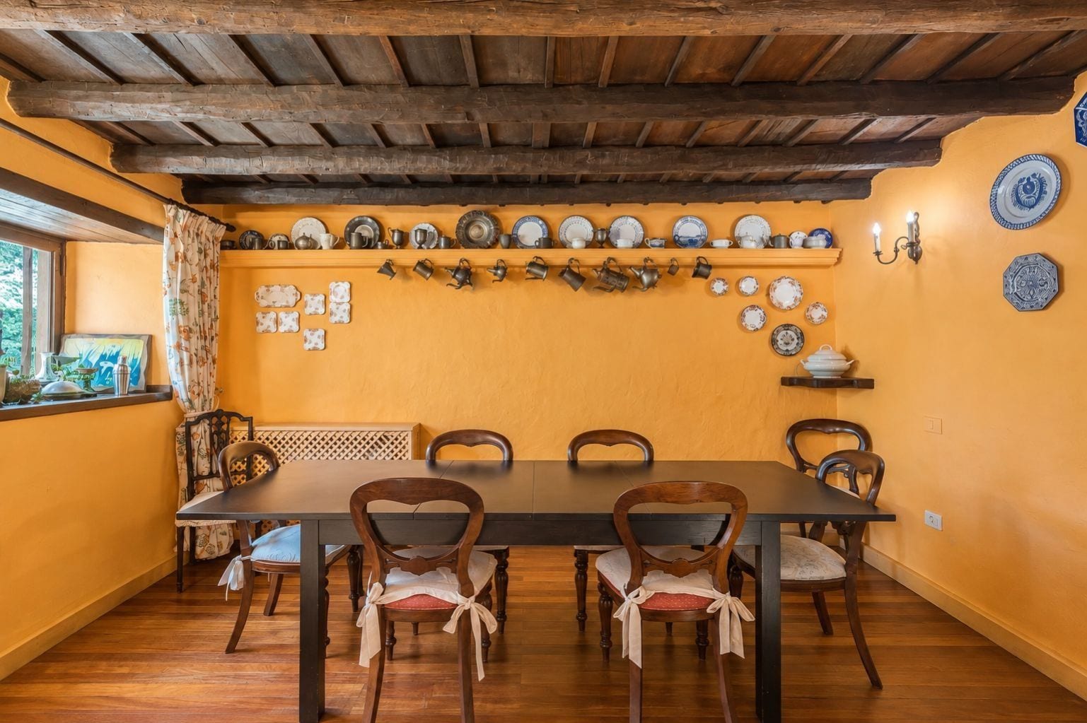 Un comedor con una mesa y sillas de madera, una pared amarilla y una variedad de platos y tazas exhibidos en estantes.
