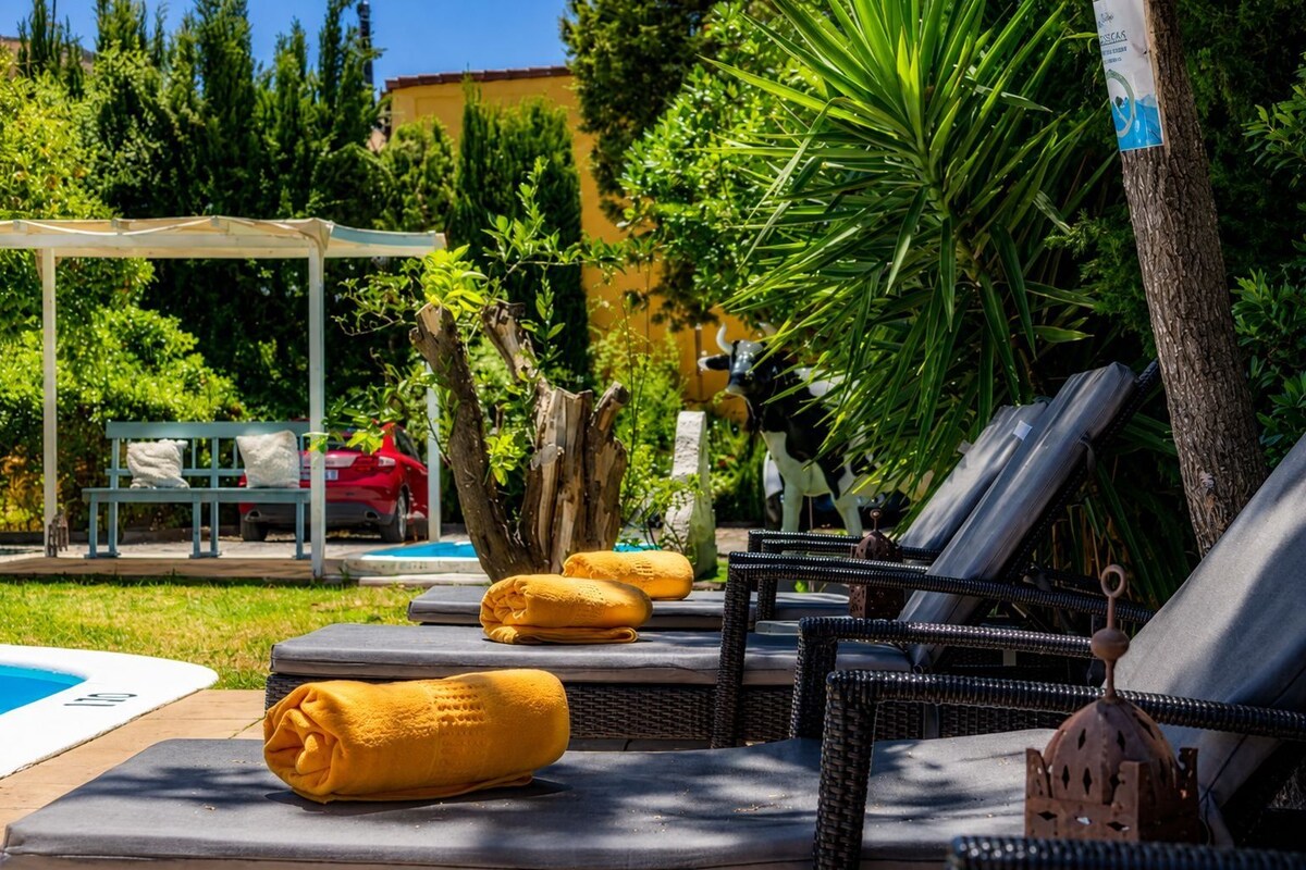 Zona junto a la piscina con sillones de descanso, un banco y una zona de estar cubierta.