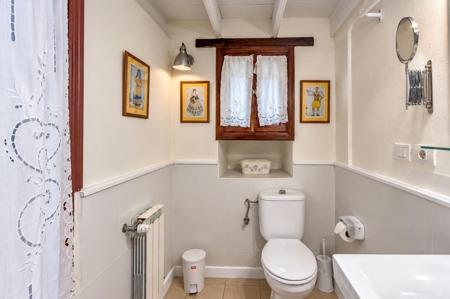 Un baño pequeño con inodoro, bidé y lavamanos. Hay cuadros en la pared y una ventana con cortinas.