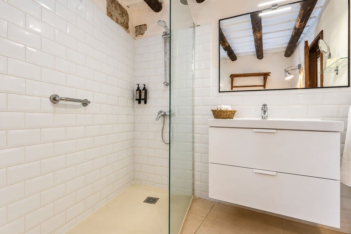 Un baño con paredes de azulejos blancos, cabina de ducha de vidrio, lavabo blanco con espejo y vigas de madera en el techo.