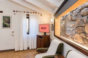 A room with a stone wall, a television displaying 'GUEST READY', a wooden cabinet, a green chair, and a window with white curtains. - Guestready - Hotel Rural Las Calas (Vega de San Mateo)