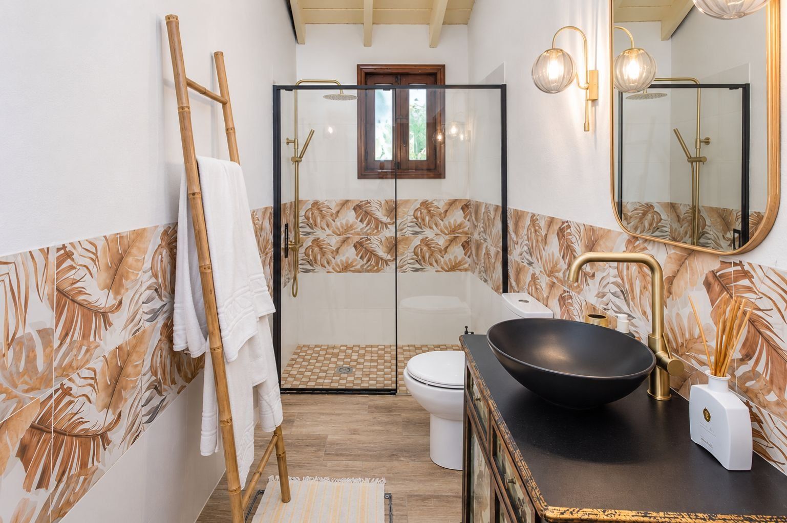 Un baño con ducha envuelta en vidrio, inodoro blanco, lavabo negro con detalles dorados y un perchero de madera.