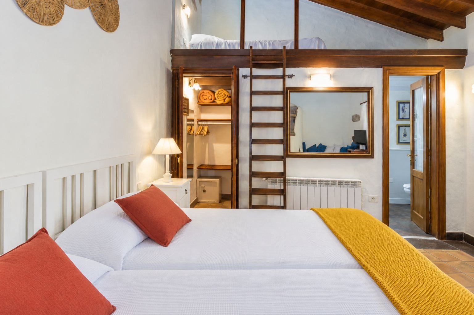 Un dormitorio con cama, almohadas, una lámpara de noche, un armario, un espejo y vista al baño.