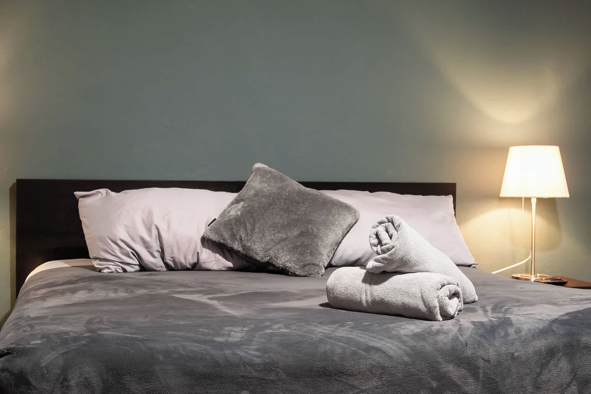Un lit bien fait, avec une couette grise, deux oreillers et une serviette pliée. Une lampe de chevet est allumée sur la droite.